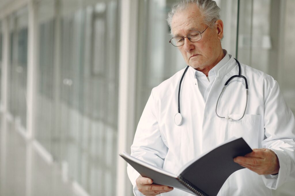 Crop concentrated professional senior male physician in white robe with stethoscope standing in modern hospital hallway and r