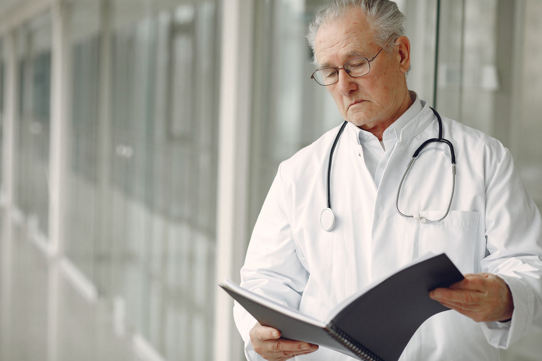 Crop concentrated professional senior male physician in white robe with stethoscope standing in modern hospital hallway and r